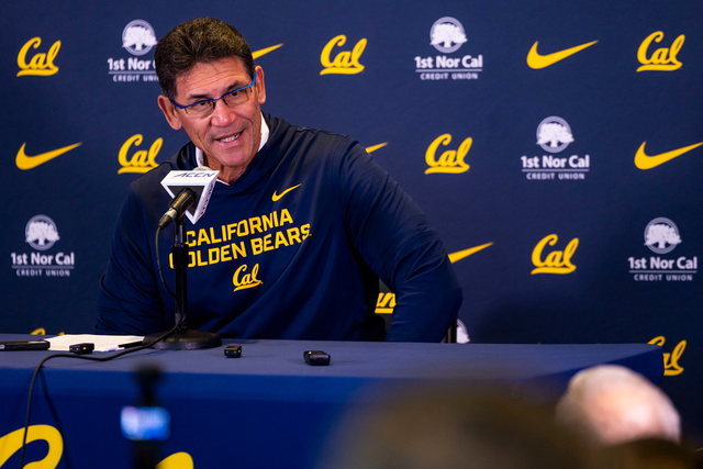 A man wearing glasses and a dark blue “California Golden Bears” sweatshirt speaks at a press conference with Cal and sponsor logos on the backdrop.